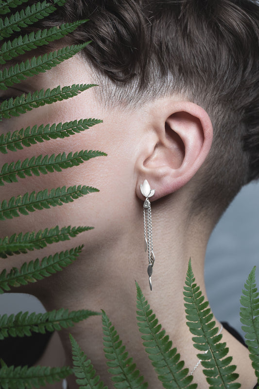 A close-up image of a person wearing a silver drop earrings, with fern leaves in the background.